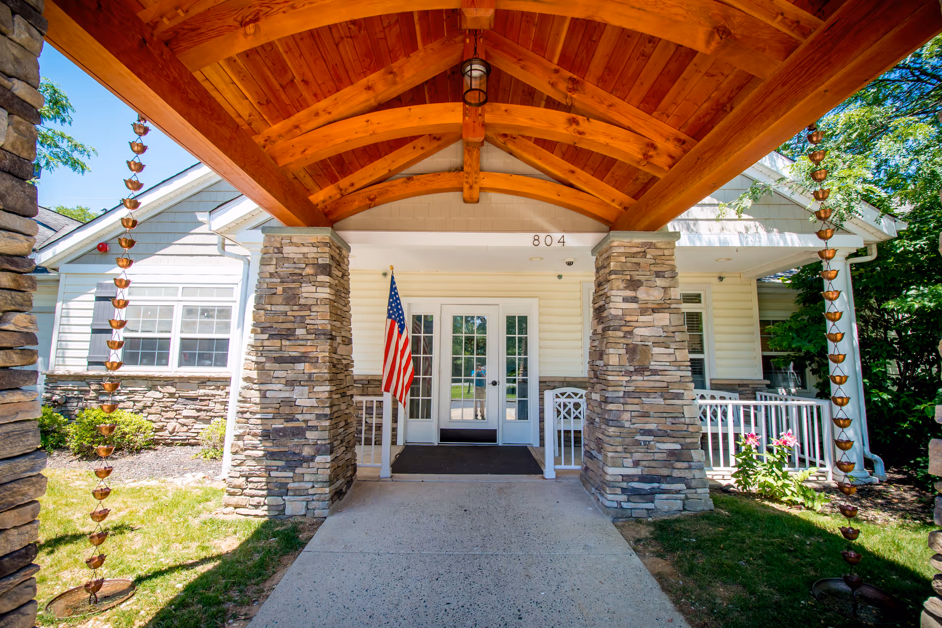 Entrance of a building with a wooden vaulted ceiling supported by stone pillars, an American flag on the left side, and double glass doors with the number 804 above them. There are plants and greenery on both sides of the entrance.