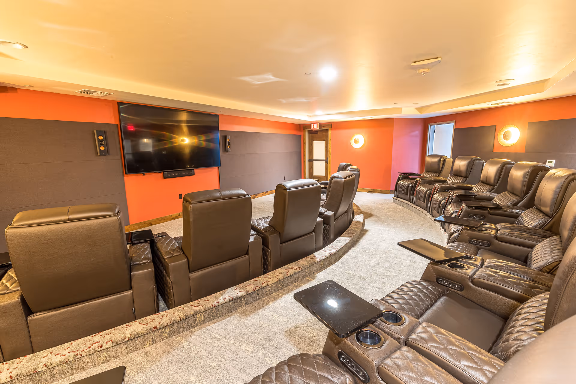 Curved screening room with rows of brown leather recliners facing a wall-mounted flat-screen TV.