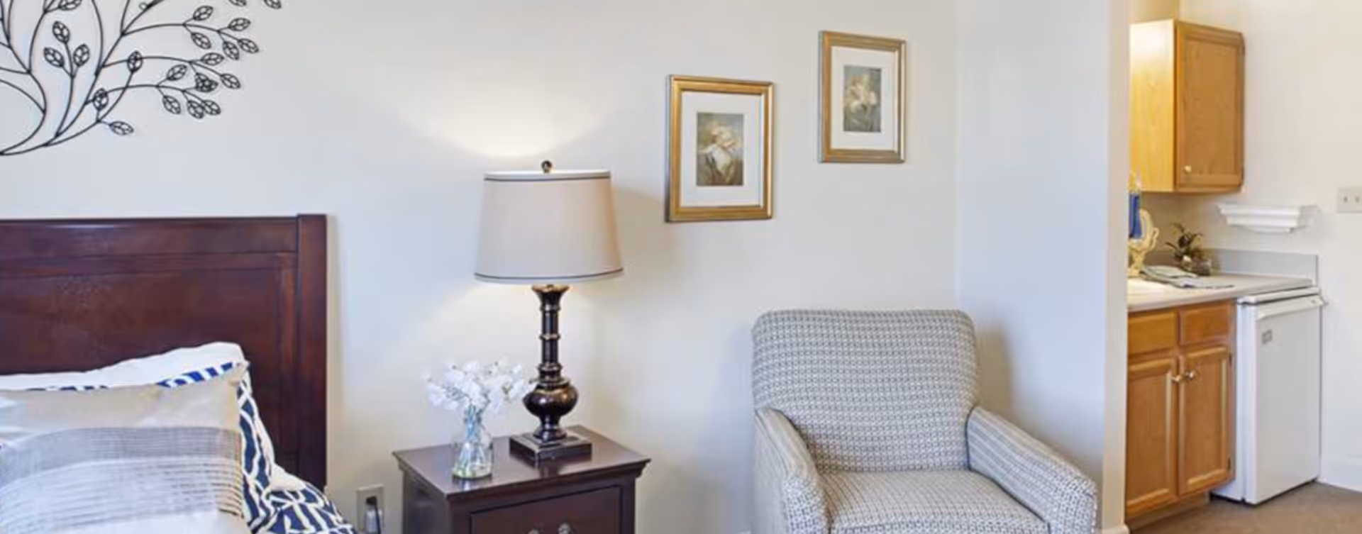 Cozy bedroom corner with a wooden headboard, nightstand and lamp, upholstered armchair, framed wall art, and a small kitchenette visible through an open doorway.