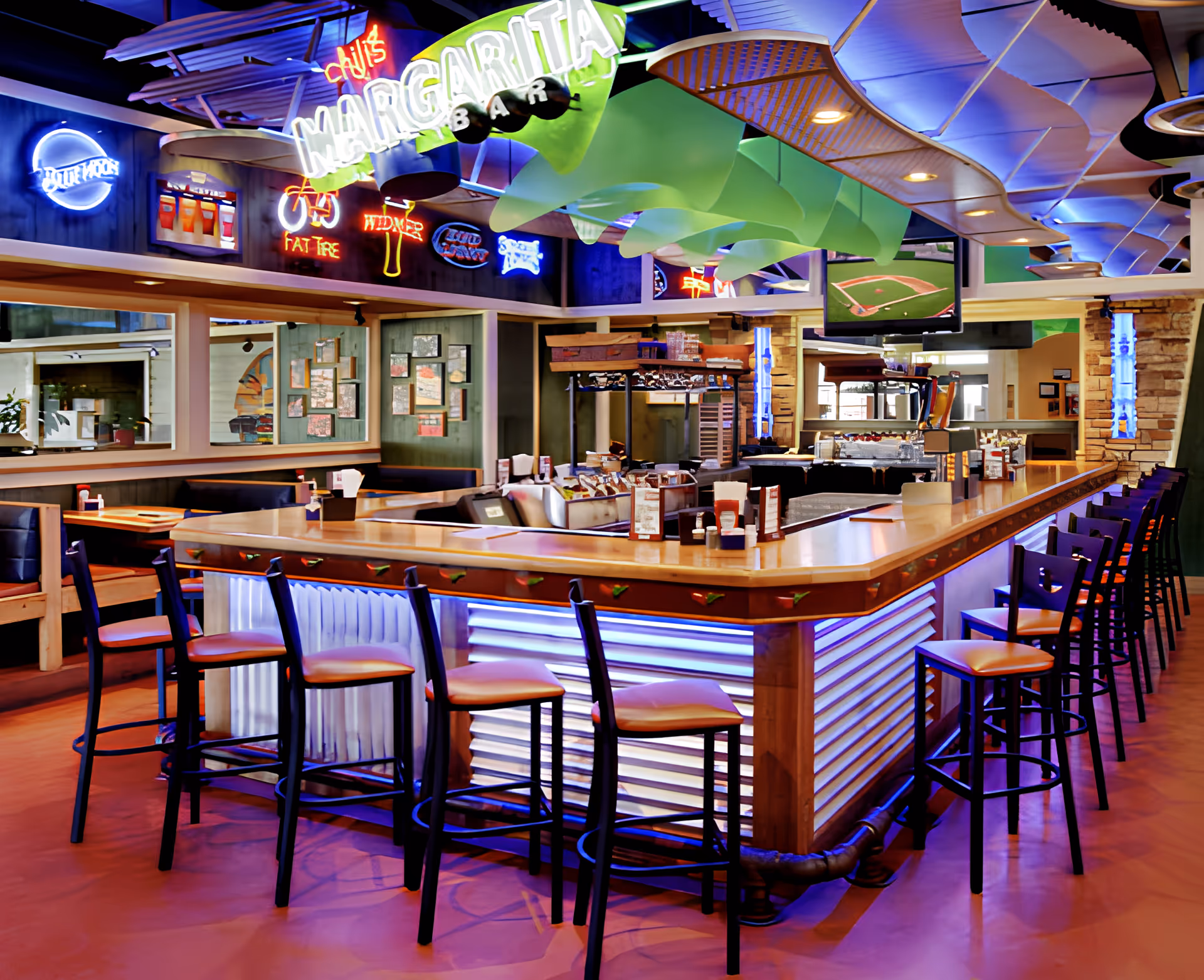 Interior view of a brightly lit bar and dining area with a long central counter, bar stools, neon signs, and overhead decorative panels.