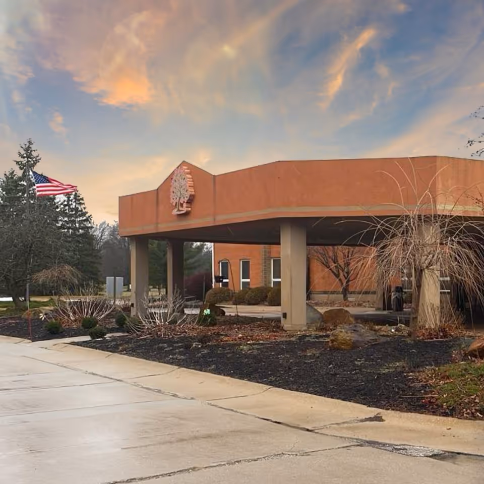 Exterior view of The Greens at Westlake facility entrance with a covered drop-off area supported by columns, landscaping with bushes and trees, and an American flag on a flagpole. The sky is partly cloudy with a warm sunset glow.