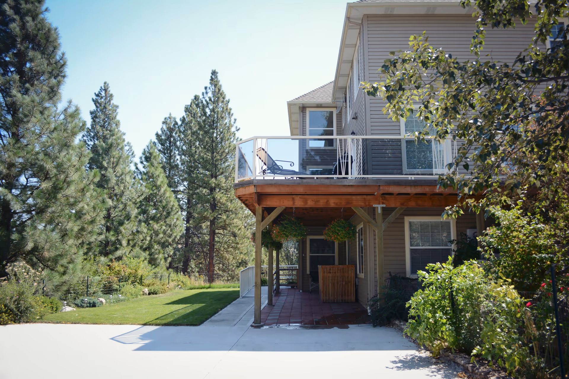 Exterior view of a two-story building with a wooden balcony on the upper floor featuring a lounge chair. The lower floor has a covered patio area with hanging flower baskets and a small wooden enclosure. Surrounding the building are green trees, bushes, and a well-maintained lawn under a clear blue sky.