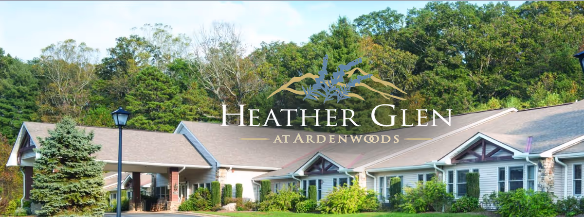 Front exterior of a single-story senior living building with landscaped shrubs and trees, overlaid with the Heather Glen at Ardenwoods logo.