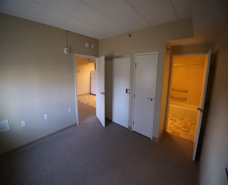 Empty room with beige walls and carpeted floor, featuring three doors: one leading to a kitchen area with a refrigerator, one to a closet, and one to a bathroom with a bathtub and grab bars.