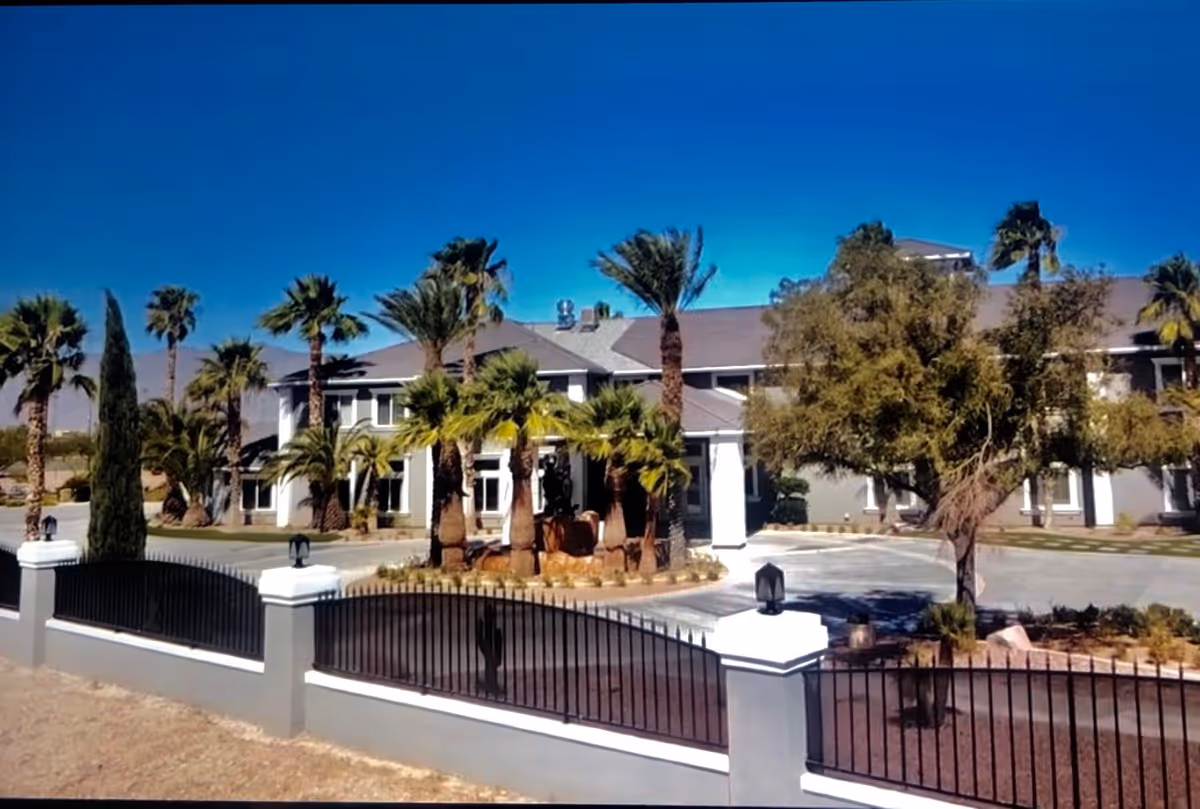 Front exterior view of a large senior living facility building with a gray roof, white pillars, and surrounded by palm trees and other greenery under a clear blue sky. A black metal fence with white pillars encloses the property.