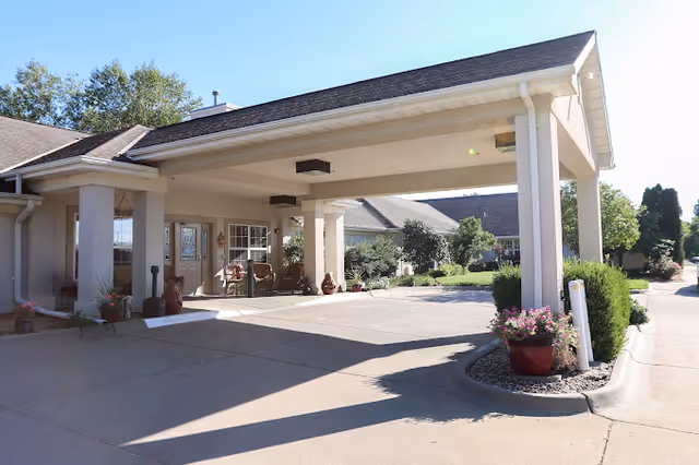 Covered porte-cochere entrance to a senior living facility with seating, potted plants, and a driveway.