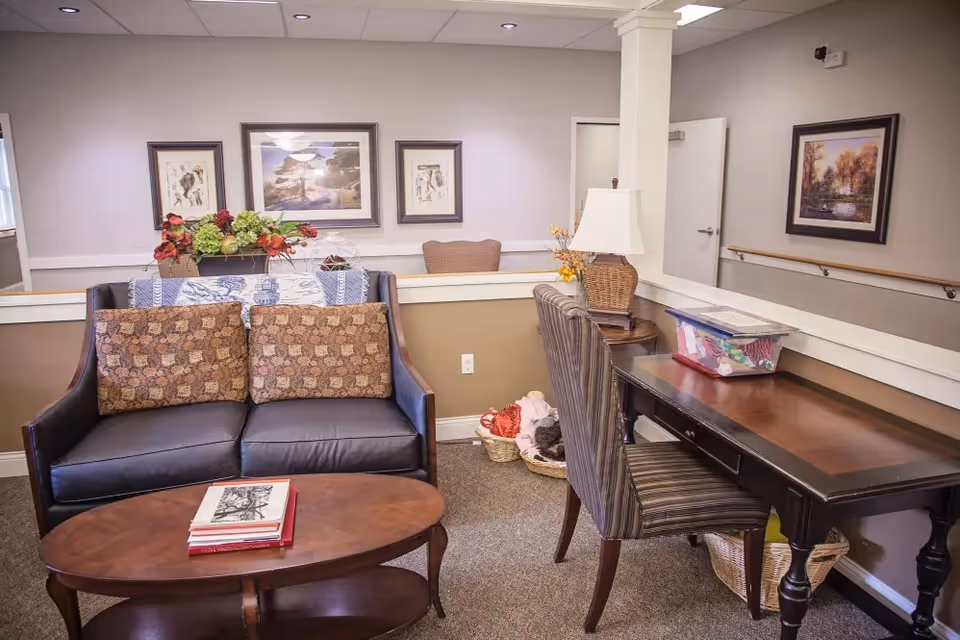 A cozy sitting area in a memory care facility featuring a dark leather loveseat with patterned cushions, a wooden coffee table with books, a wooden desk with a striped chair, a table lamp, and decorative framed artwork on the walls. There are baskets with items on the floor and a partial view of a hallway with a handrail.