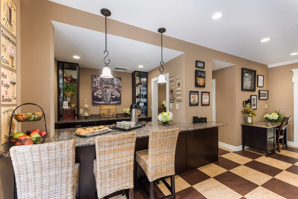 Interior view of a kitchen area with a granite countertop island featuring three wicker bar stools. The countertop has a tray with pastries, a coffee pot, and cups. The walls are decorated with vintage-style coffee and dessert signs, and there are pendant lights hanging above the island. In the background, there are shelves with decorative items and a framed picture of a classic car. To the right, there is a small desk with a chair and floral arrangements on top. The floor has a checkered pattern in brown and beige tones.