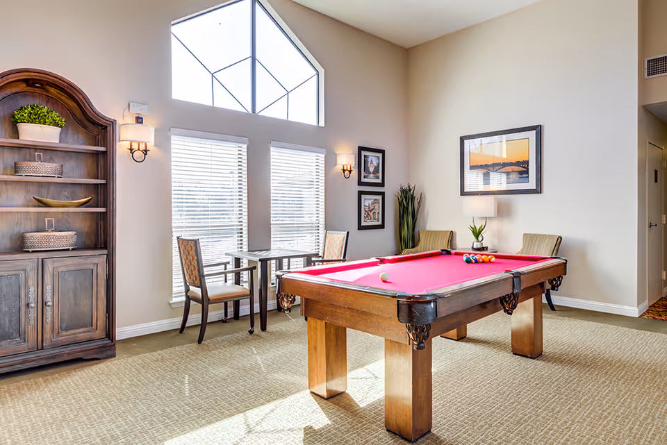 A bright room with a red felt pool table in the center, surrounded by chairs and a small table near large windows with blinds. The room has beige walls, a high triangular window, a wooden cabinet with decorative items, framed pictures on the walls, and a lamp on a side table.