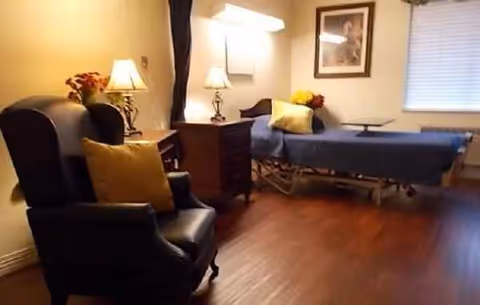 A cozy patient room with a blue hospital bed, chair, lamps, and framed art.