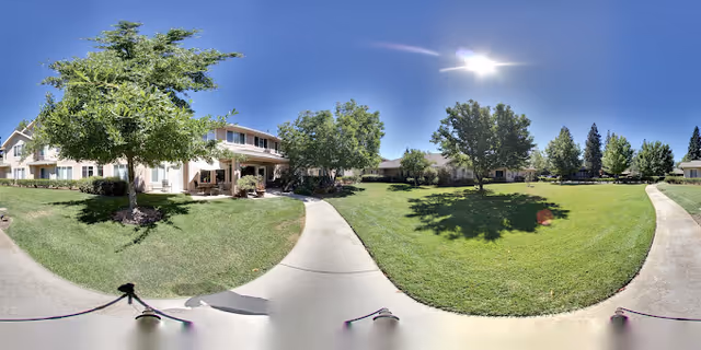 A sunny outdoor scene at Marbella Chico featuring a well-maintained grassy area with several trees and a paved walkway. Residential buildings with light-colored walls and multiple windows surround the green space under a clear blue sky.