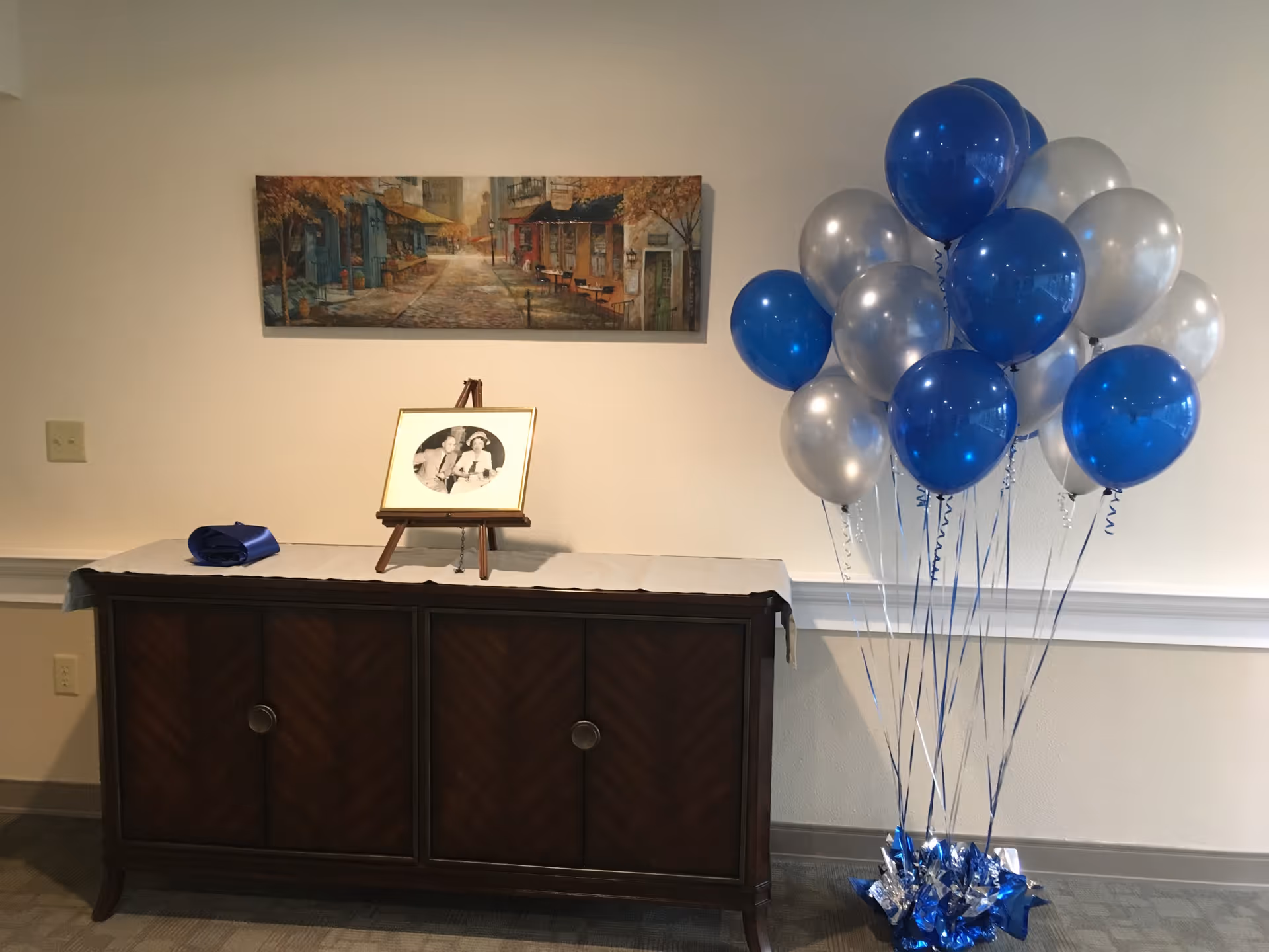 A wooden cabinet with a white runner on top, holding a framed black and white photo on a small easel. To the right of the cabinet is a bunch of blue and silver balloons tied together. Above the cabinet hangs a painting of a street scene with buildings and trees.