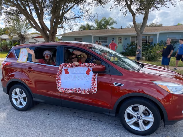A red SUV decorated with holiday signs and garland carries people past a single-story house during an outdoor parade.