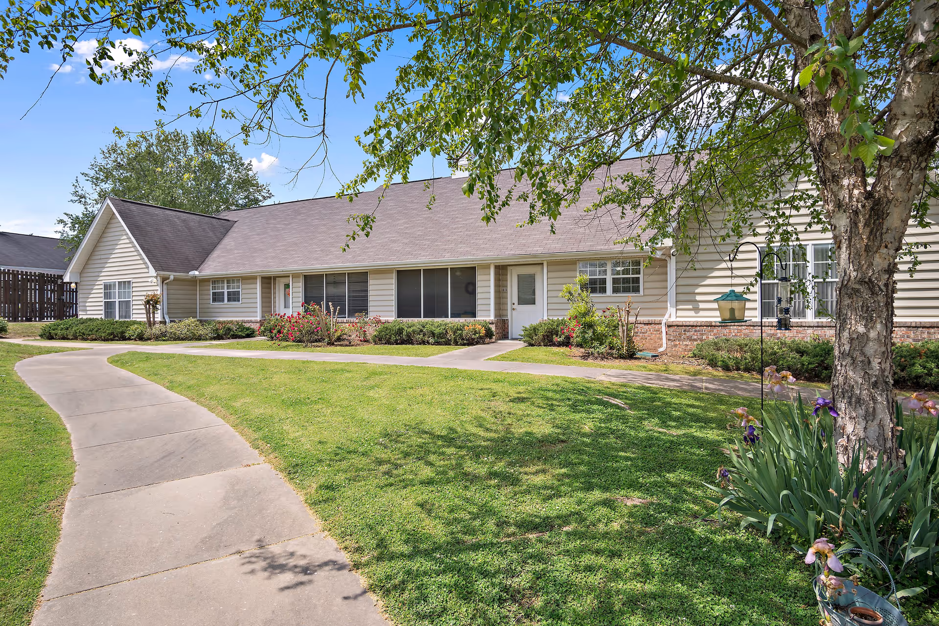 Exterior view of a single-story residential building with beige siding and a brown roof, surrounded by a well-maintained lawn, shrubs, and flowers. A curved concrete walkway leads to the front doors. A tree with green leaves and flowering plants is visible in the foreground under a clear blue sky.
