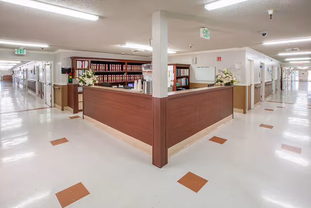 Interior view of a senior living facility hallway with a central wooden reception desk. The hallway is well-lit with white walls and ceiling, and the floor has a white and brown tile pattern. There are exit signs and fire safety equipment visible on the walls.