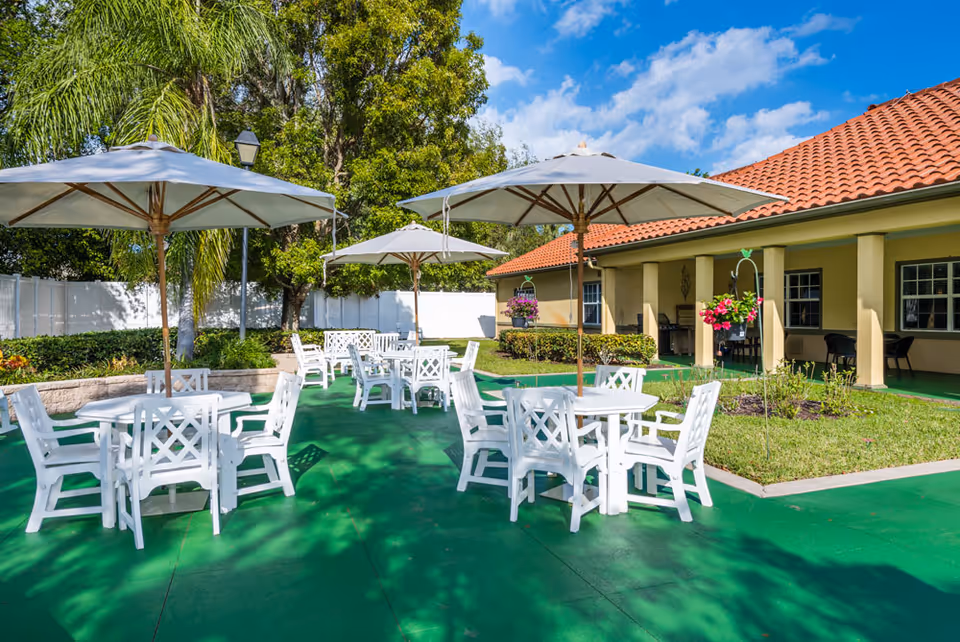 Outdoor patio area with white tables and chairs under large white umbrellas, surrounded by greenery and a building with a red-tiled roof in the background under a blue sky with some clouds.