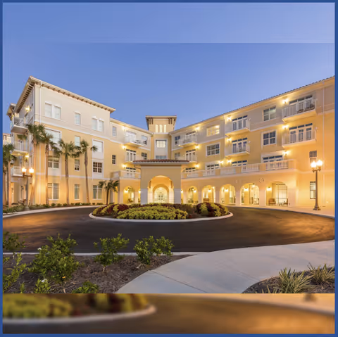 Exterior view of a large, well-lit senior living facility building at dusk with multiple floors, balconies, palm trees, and a circular driveway with landscaped plants in the center.