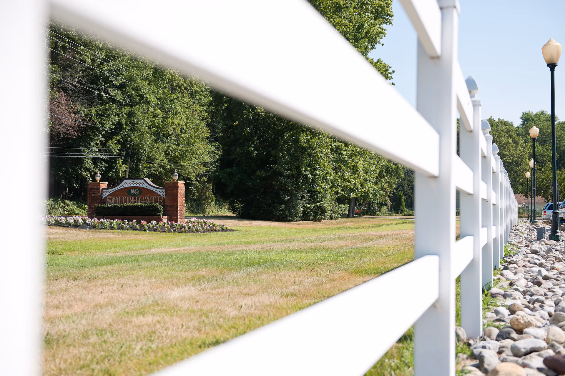 View of a white wooden fence running alongside a grassy area with a landscaped flower bed and a brick sign that reads 'Southgate'. Trees and lamp posts are visible in the background under a clear sky.
