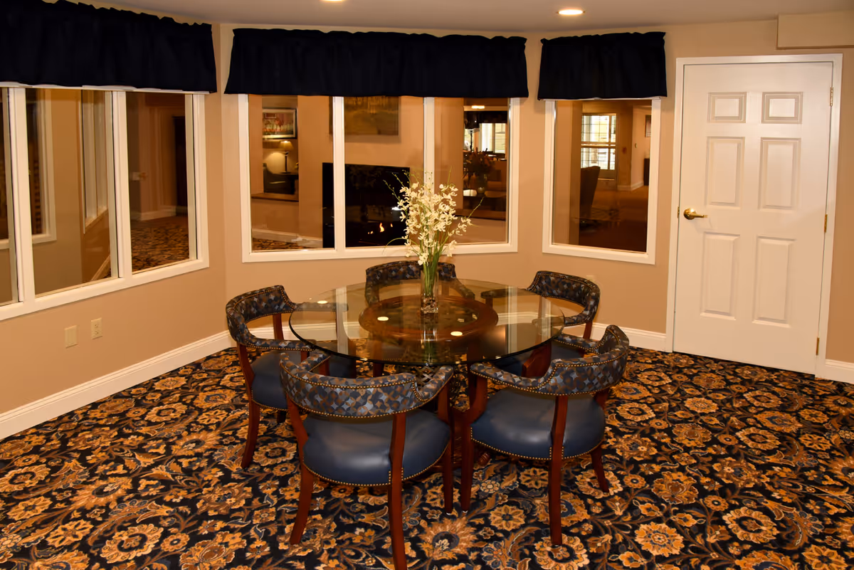 Carpeted dining area with a round glass table surrounded by six upholstered chairs, a floral centerpiece, windows, and a door.
