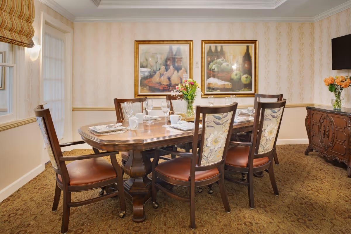 A formal dining room with a wooden table set for six, upholstered chairs, a floral centerpiece, and framed paintings on the wall.
