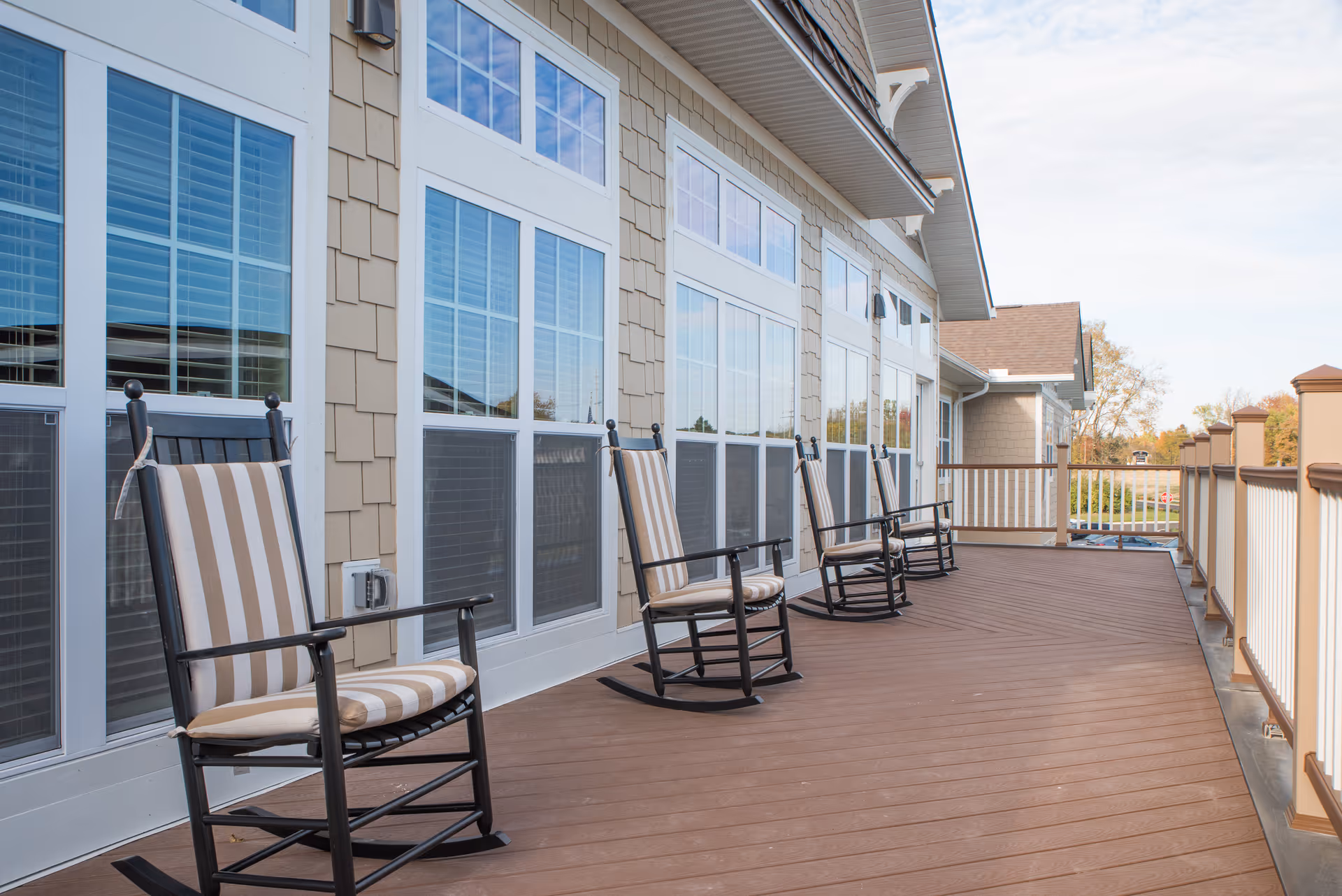 A spacious outdoor deck area with four black rocking chairs featuring beige and white striped cushions. The deck has brown wooden flooring and white railings with brown posts. Large windows with white frames line the building wall adjacent to the deck, reflecting the blue sky and surrounding scenery.