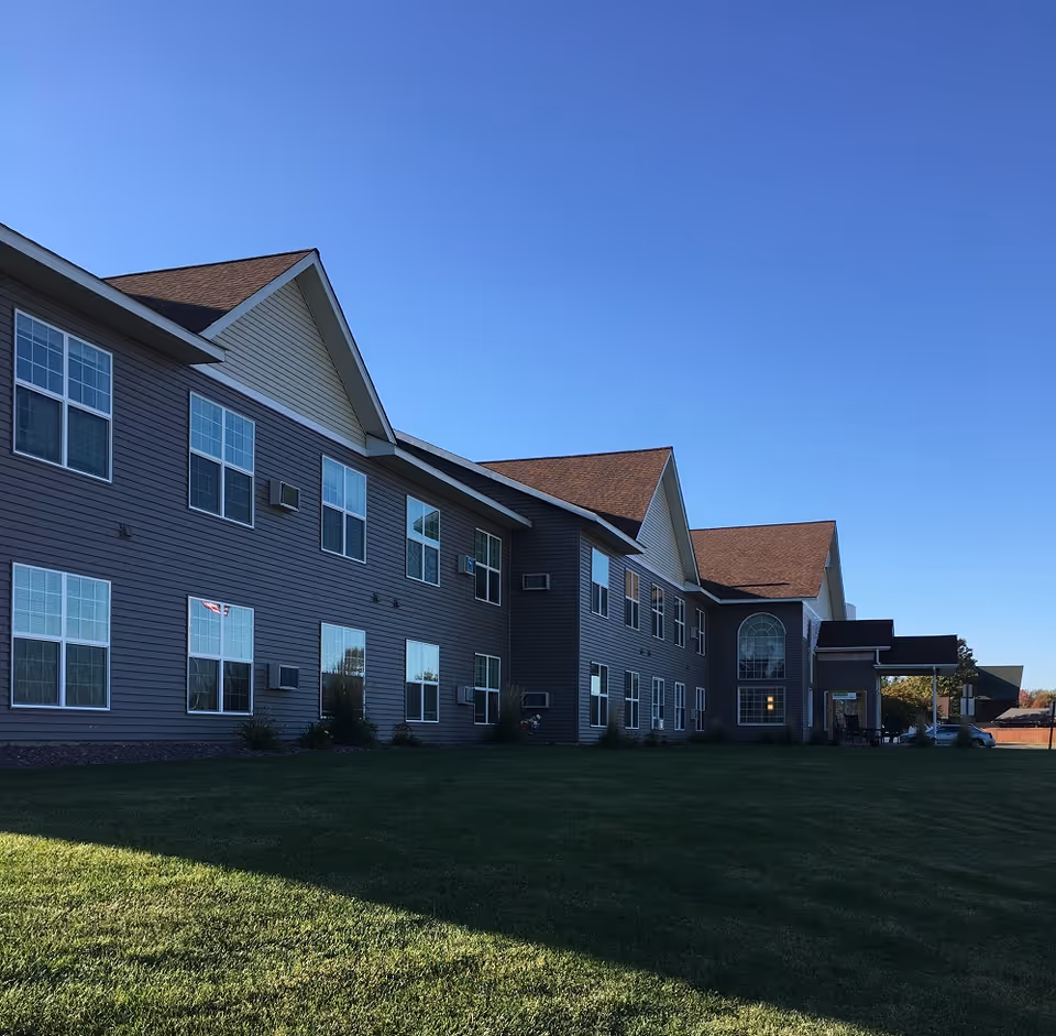 Exterior view of a two-story senior living facility building with multiple windows, a sloped roof, and a well-maintained green lawn under a clear blue sky.