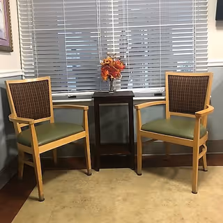 Two wooden chairs with green cushions and woven backs are placed on either side of a small dark wooden table with a vase of orange and red flowers. Behind them is a window with closed white blinds.