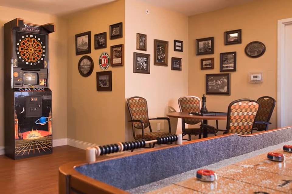 A recreational room with a dartboard game machine on the left, a round table with four patterned chairs, and framed pictures on beige walls. In the foreground, there is a shuffleboard table with pucks on it.