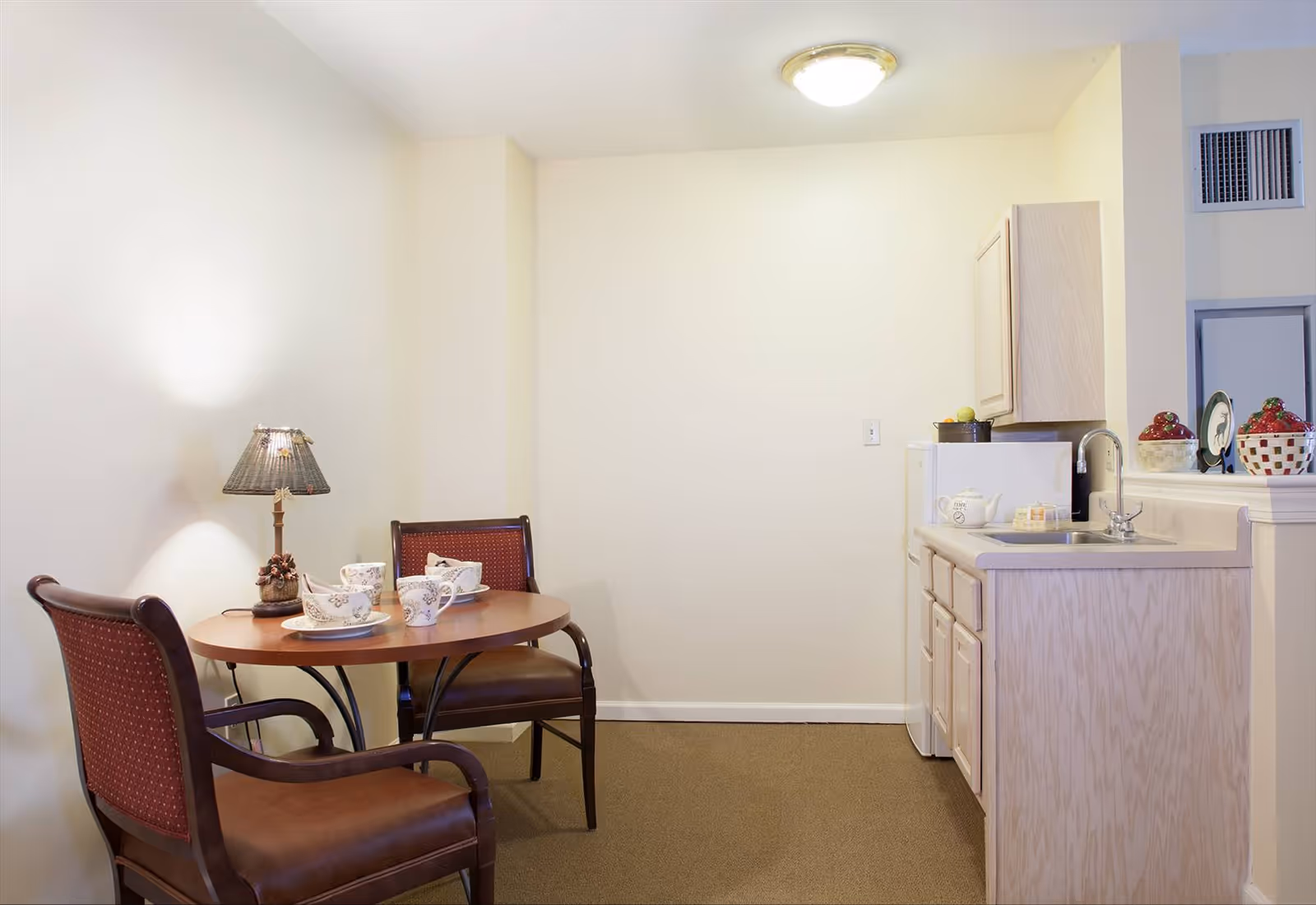 Small dining nook with a round table set for tea, two upholstered chairs, a lamp, and a compact kitchenette with sink and cabinets.