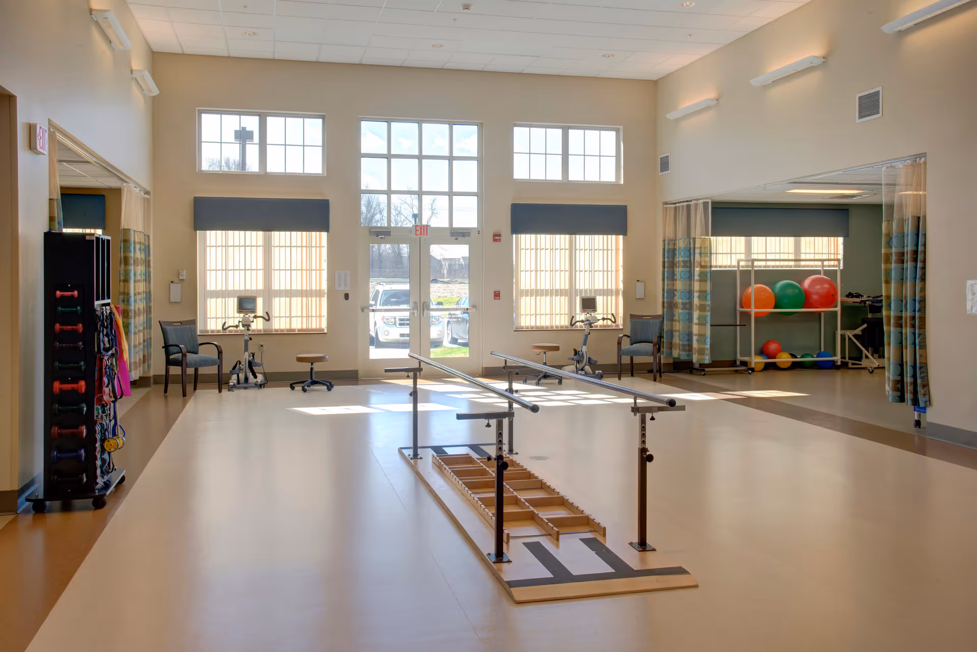 A spacious rehabilitation or physical therapy room with parallel bars in the center for walking exercises. The room has large windows and a glass door letting in natural light. There are exercise balls on a rack, stationary bikes, chairs, and various exercise equipment along the walls. The floor is smooth and suitable for mobility exercises.