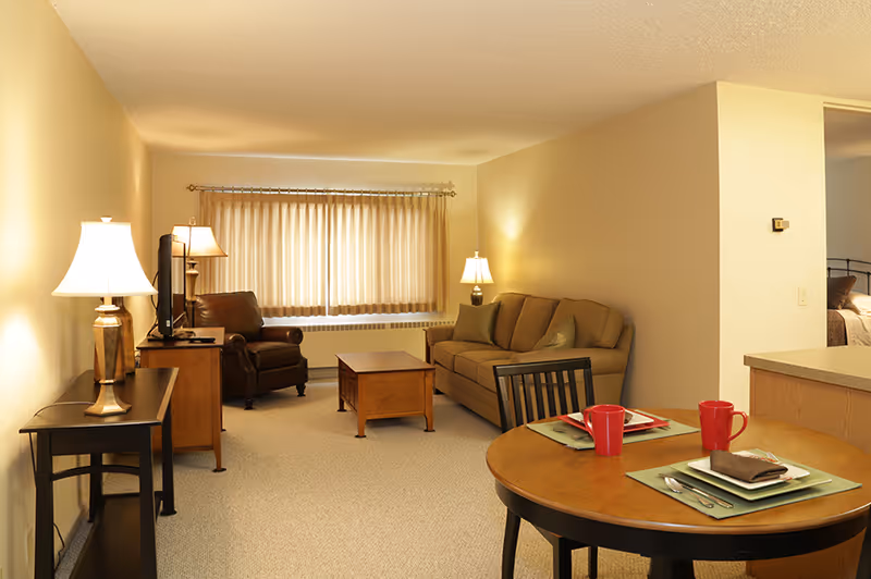 A cozy living room area in a senior living facility with beige walls and carpet. The room features a brown sofa with cushions, a wooden coffee table, a leather armchair, and two table lamps providing warm lighting. A flat-screen TV is placed on a wooden stand near the armchair. In the foreground, there is a round wooden dining table set for two with red mugs, plates, and silverware. A window with beige curtains is visible in the background, and a partial view of a bedroom is seen through an open doorway.