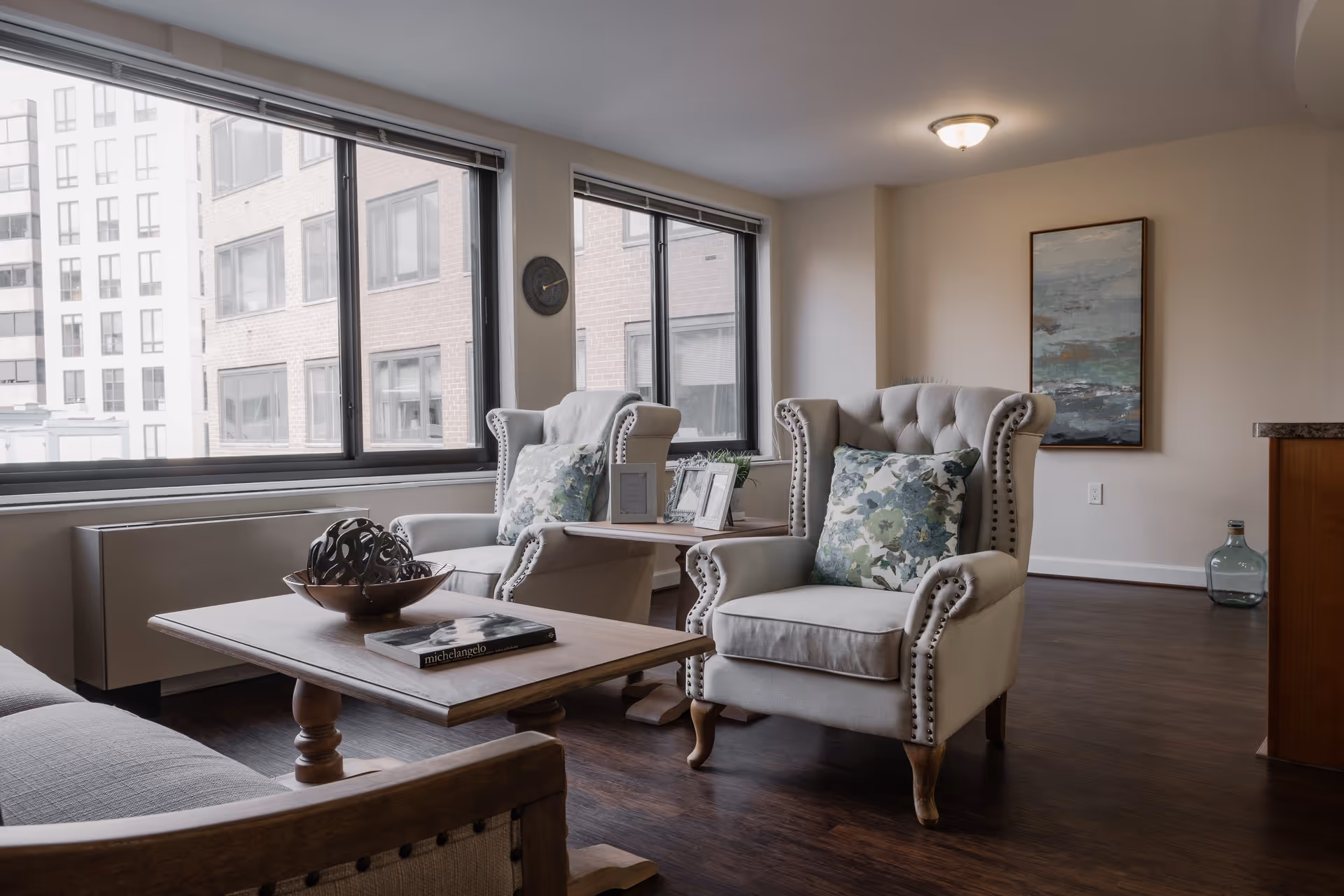 A cozy living room area with two beige armchairs featuring floral cushions, a wooden coffee table with a decorative bowl and a book, large windows showing an urban building exterior, and a framed abstract painting on the wall.