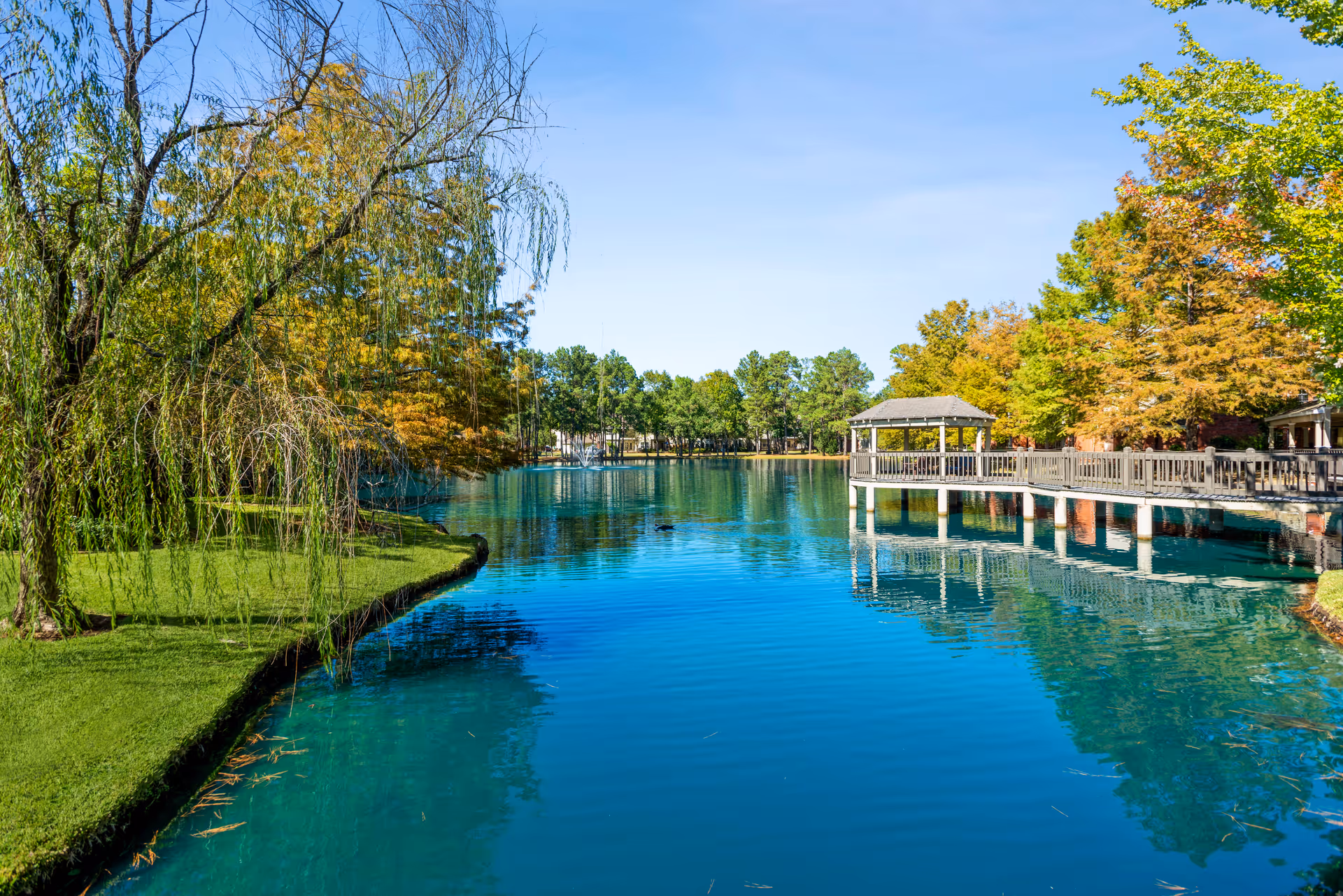 A serene outdoor scene featuring a clear blue pond surrounded by lush green grass and trees with autumn foliage. A wooden pier with a gazebo extends over the water on the right side, under a clear blue sky.