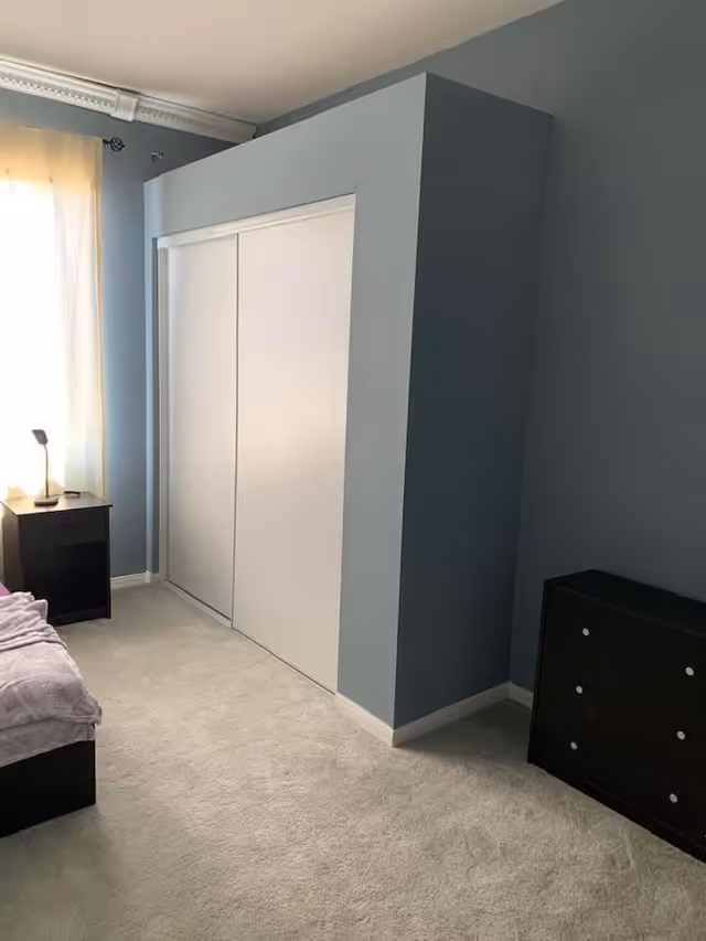 Carpeted bedroom corner with a white sliding-door closet, nightstand with a lamp, dresser, and part of a bed visible.