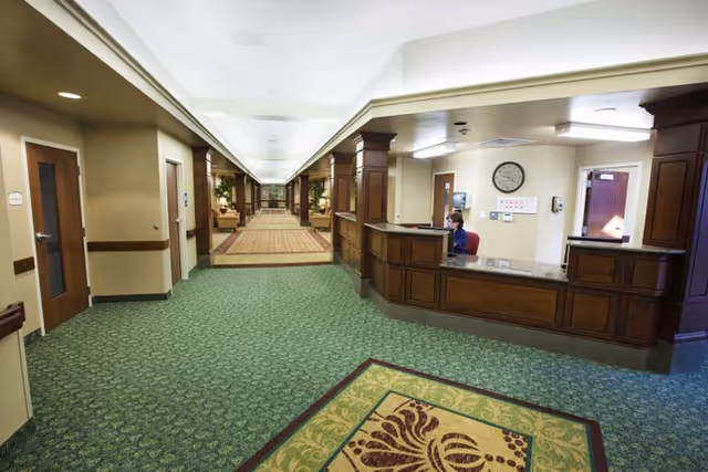 Interior view of a senior living facility hallway with green patterned carpet and beige walls. On the right side, there is a wooden reception desk with a person seated behind it. The hallway extends into the distance with seating areas and plants visible along the corridor.