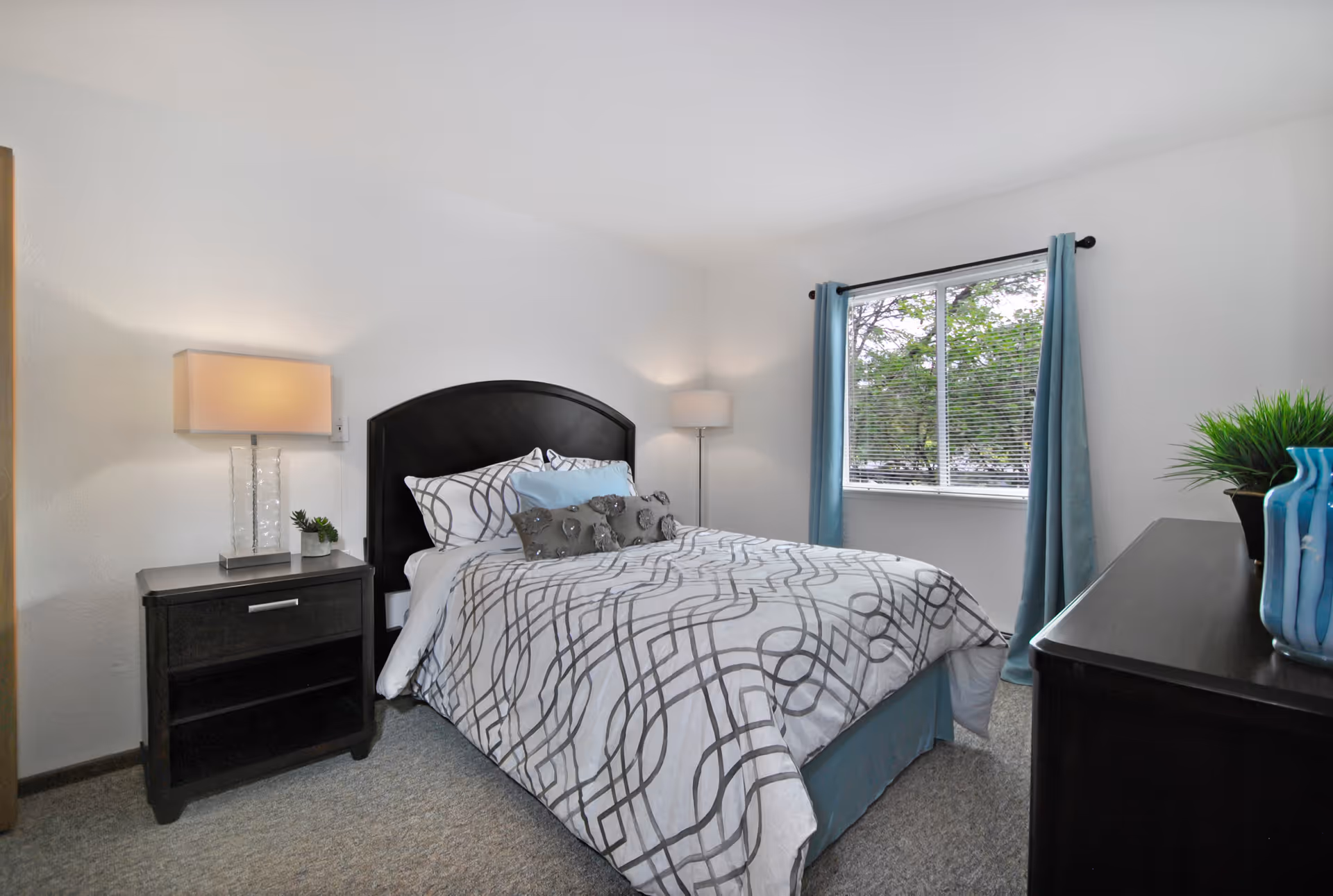 A neatly made bed with patterned white and gray bedding and multiple pillows in a bedroom. There is a dark wooden nightstand with a lamp and small plant on the left side of the bed. A window with blue curtains lets in natural light, and a dark wooden dresser with decorative vases and a plant is on the right side of the room. The walls are white and the floor is carpeted.