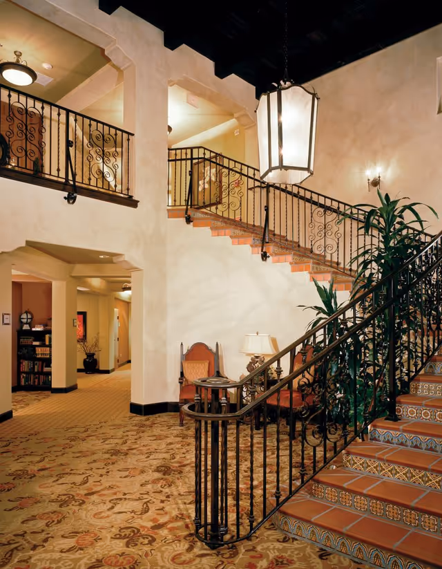 Interior view of a senior living facility featuring a decorative staircase with wrought iron railings and patterned tile steps. The area includes a carpeted floor with floral designs, a large hanging lantern light fixture, a small seating area with chairs and a table lamp, and potted plants. The walls are painted in warm tones with archways leading to other rooms.