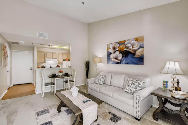 A cozy living room with a light gray sofa adorned with two patterned pillows, a wooden coffee table with a decorative runner and a white vase, and a side table with a lamp and some small items. Behind the sofa is a large floral painting. To the left, there is a small dining area with a round table set for two and a view into a kitchen with light wood cabinets and white appliances. The walls are painted a soft beige color.