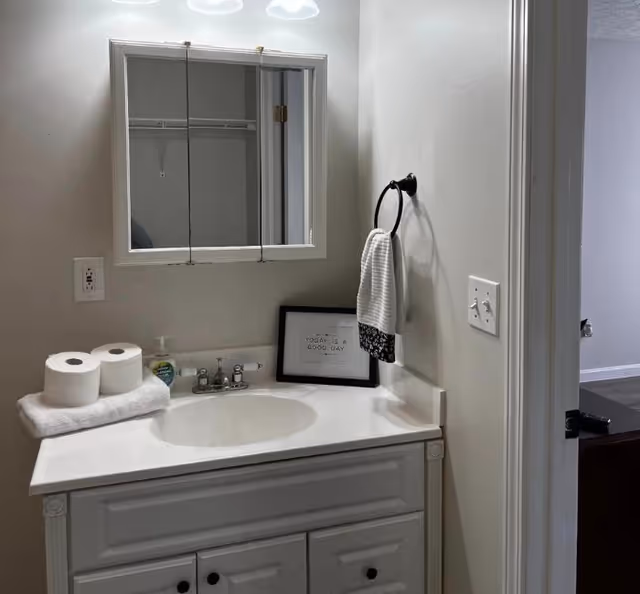 White bathroom vanity with sink and mirror cabinet, hand towel on a ring, soap dispenser and two rolls of toilet paper on the counter.