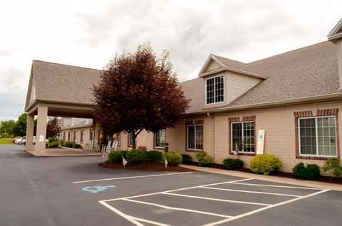 Exterior view of a single-story brick building with a covered entrance, several windows, and a parking lot with marked handicap spaces. There are small bushes and a tree near the building, under a cloudy sky.