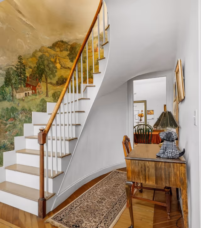 Interior view of a senior living facility hallway featuring a curved staircase with wooden handrail and white steps. The wall beside the staircase has a mural depicting a countryside scene with trees, houses, and hills. A wooden table with a lamp and a decorative dog figurine is placed along the wall, and a patterned rug runs along the wooden floor. In the background, a dining area with a table and chairs is visible through a doorway.