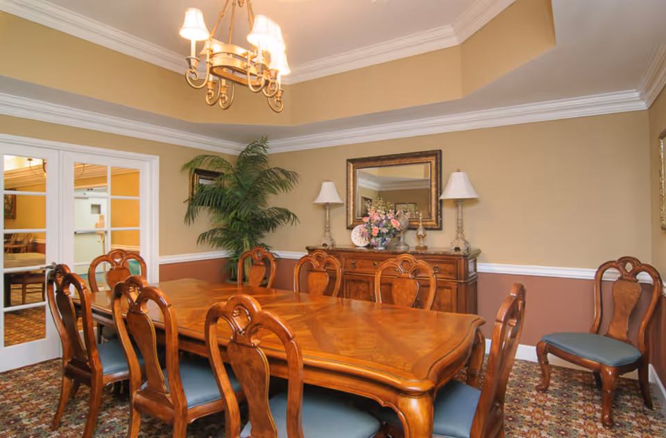 A formal dining room with a large wooden dining table surrounded by eight matching wooden chairs with blue cushions. The room features beige walls with white crown molding and a two-tone paint design. A wooden sideboard with two lamps, a floral arrangement, and a framed mirror is against one wall. A potted plant is in the corner, and a chandelier with five lights hangs above the table. French doors with glass panes lead to another room.