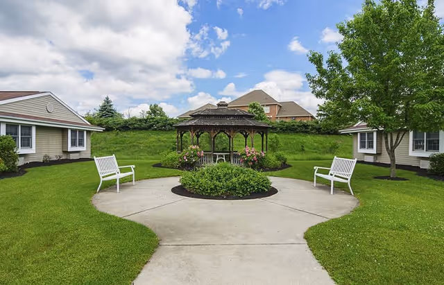 Outdoor courtyard area with a central gazebo surrounded by green grass and bushes. There are white benches on either side of a concrete pathway leading to the gazebo. Two single-story buildings with beige siding and white trim flank the courtyard. A large tree is visible on the right side, and a partly cloudy blue sky is overhead.
