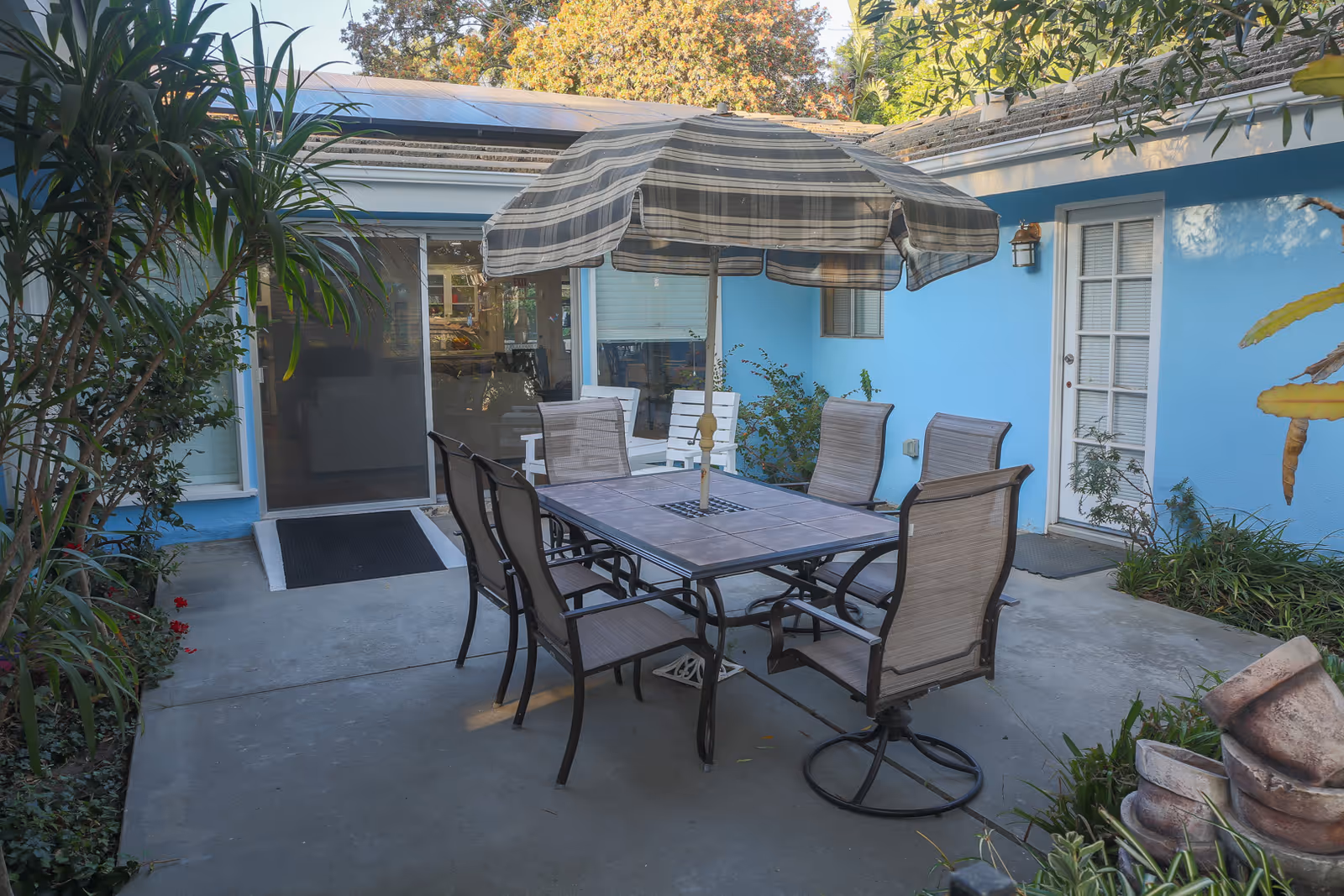 Outdoor patio area with a rectangular table and six chairs around it, shaded by a large striped umbrella. The patio is surrounded by blue walls with windows and doors, some plants, and trees visible in the background.