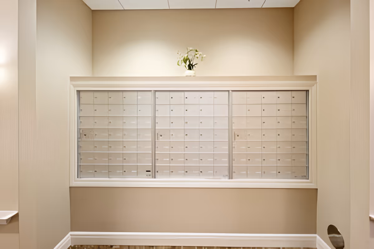 A beige-colored mailroom wall with a large set of locked mailboxes behind glass panels. Above the mailboxes is a small vase with green and white flowers. The room has soft lighting and a clean, simple design.