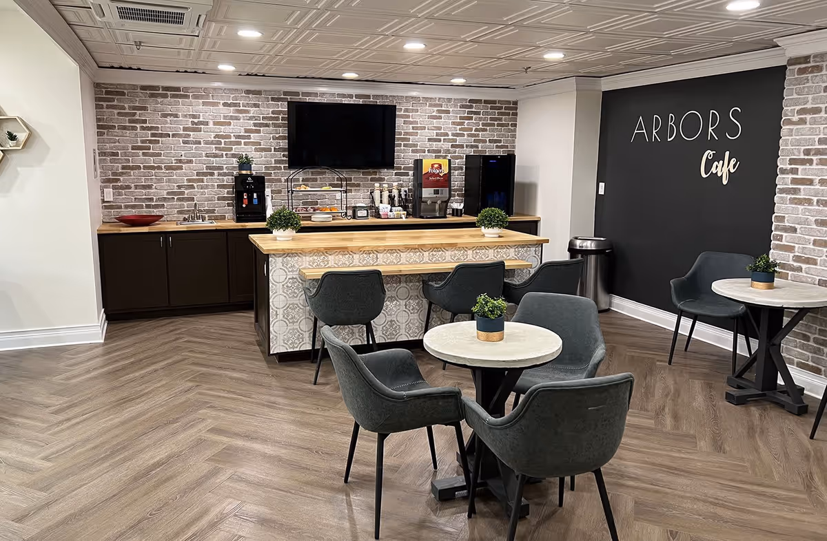 A cozy cafe area inside The Arbors facility featuring a small counter with a coffee machine, a beverage dispenser, and a TV mounted on a brick wall. There are several round tables with gray upholstered chairs and small potted plants on each table. The wall has a blackboard with the words 'ARBORS Cafe' written on it. The floor has a wood-patterned design.