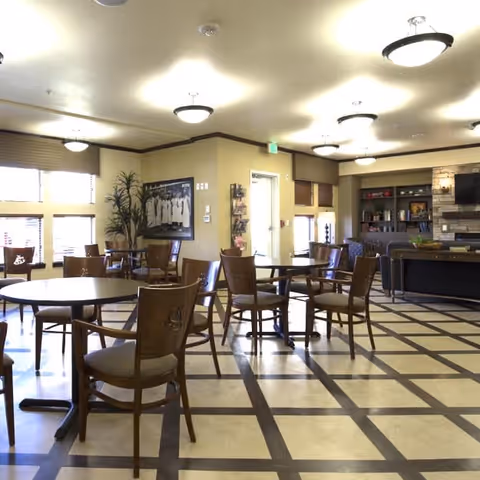 A spacious common area with several round tables and wooden chairs arranged on a checkered floor. The room has large windows with blinds, a few plants, framed artwork on the walls, and built-in shelves with decorative items. Ceiling lights provide illumination, and there is a stone fireplace with a mounted TV above it in the background.