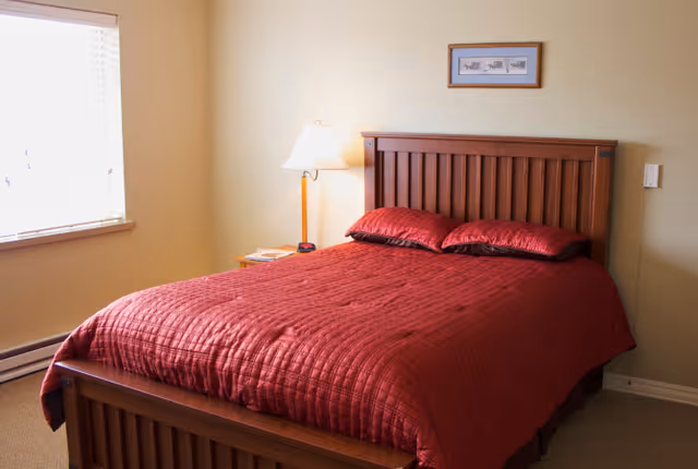 A bedroom with a wooden bed frame and headboard, a red quilted bedspread with matching pillows, a bedside table with a lamp and some items on it, a window with blinds, and a framed picture on the wall above the bed.