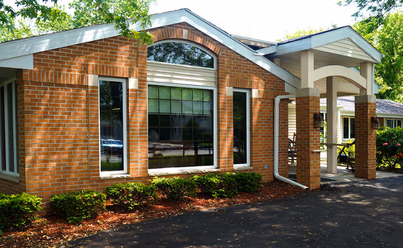 Brick building exterior with large front windows, entry porch, and landscaped shrubs.