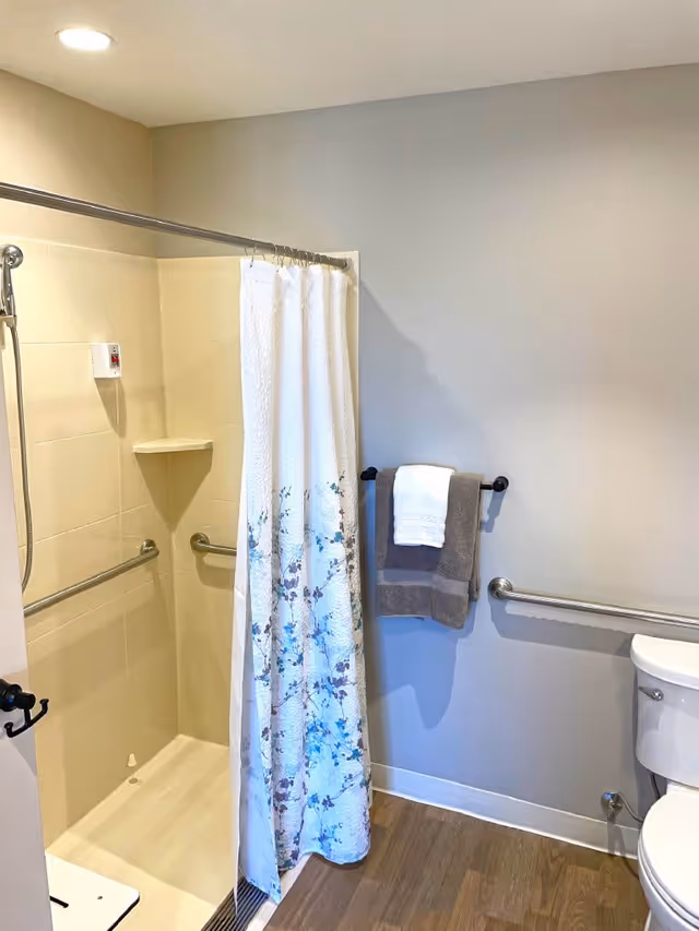 A bathroom with a walk-in shower featuring beige tiles, a white shower curtain with a blue floral pattern, and stainless steel grab bars. There is a towel rack on the wall holding a gray towel with a white towel on top. The floor is wood-style, and a white toilet is partially visible on the right side.